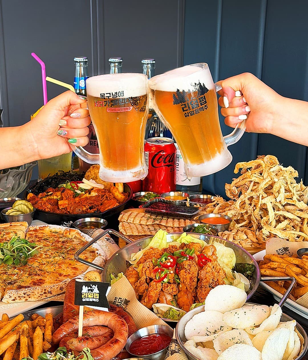 강원특별자치도 춘천시 리얼펍 춘천온의점 맥주,호프 - 살얼음 맥주잔을 맞댄 상황과 치킨, 소시지, 피자가 가득한 테이블. 1인 좌석 이용이 가능한 안주 구성과 조명 아래 맥주 거품의 질감.