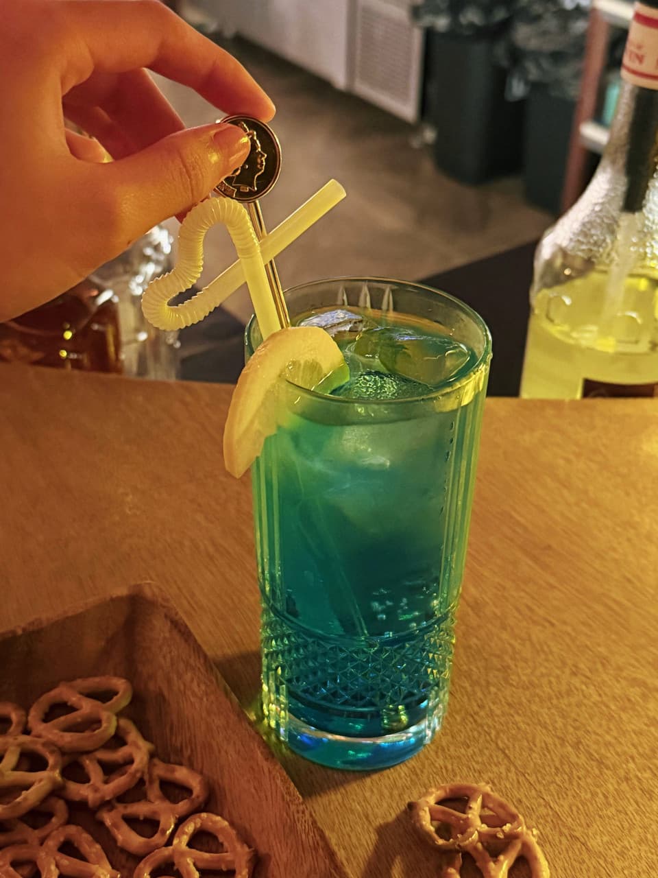 강원특별자치도 강릉시 난장혼술바 강릉점 바(Bar) 조명 아래 파란색 칵테일과 프레첼. 나무 다찌석에서 하트 빨대가 꽂힌 잔을 든 1인 혼술 상황.