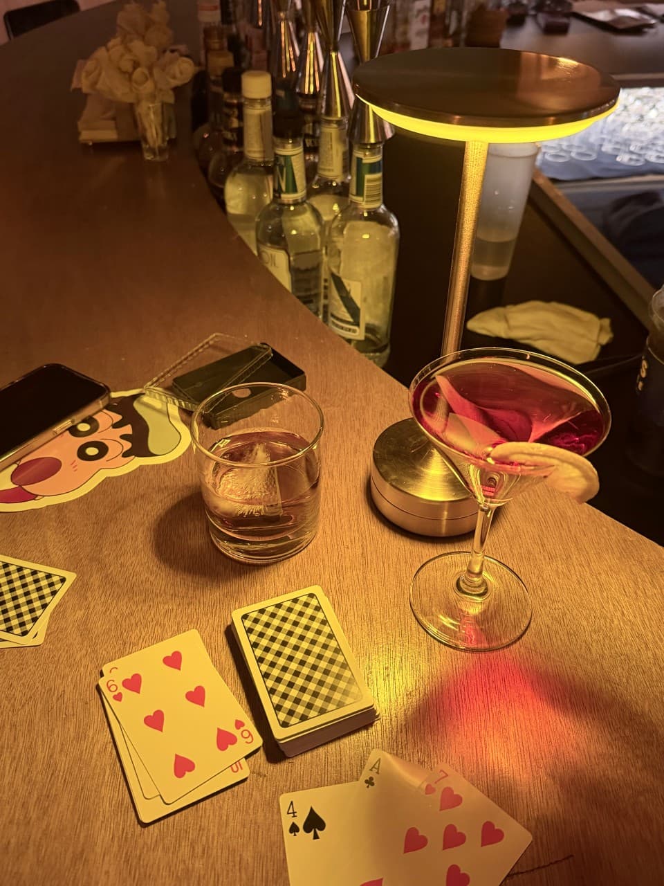강원특별자치도 강릉시 난장혼술바 강릉점 바(Bar)의 노란 조명이 비치는 1인 다찌석. 목재 테이블에 놓인 붉은 칵테일과 위스키, 트럼프 카드로 구성된 혼술 상황.