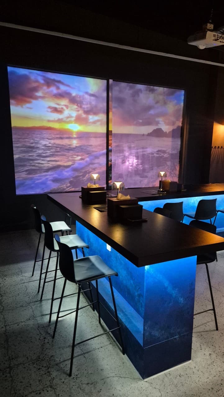 강원특별자치도 강릉시 혼술바 파도끝 바(Bar) 내부. 바다 노을 영상이 투사된 벽면 앞 푸른 조명의 다찌석과 1인용 의자가 배치되어 혼술이 가능한 카운터 테이블 상황.