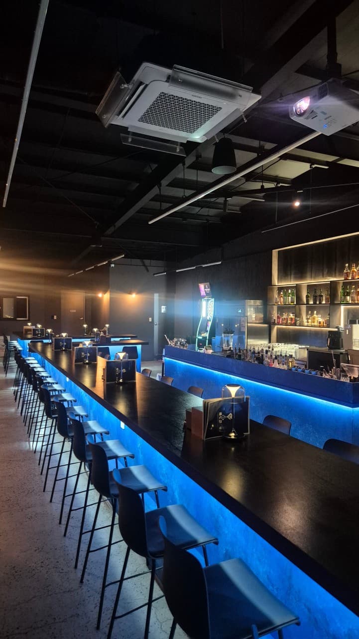 강원특별자치도 강릉시 혼술바 파도끝 바(Bar) 푸른 조명이 비치는 긴 다찌석과 1인용 좌석. 벽면의 술병들과 다트 게임기가 배치된 실내 공간.