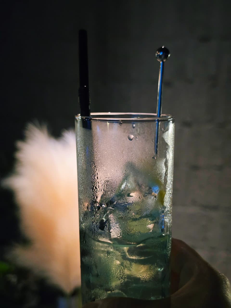강원특별자치도 강릉시 혼술바 파도끝 바(Bar) 다찌석에서 물방울 맺힌 하이볼 잔을 든 손. 어두운 조명 아래 빨대와 머들러가 꽂힌 1인 음료의 질감을 묘사함.