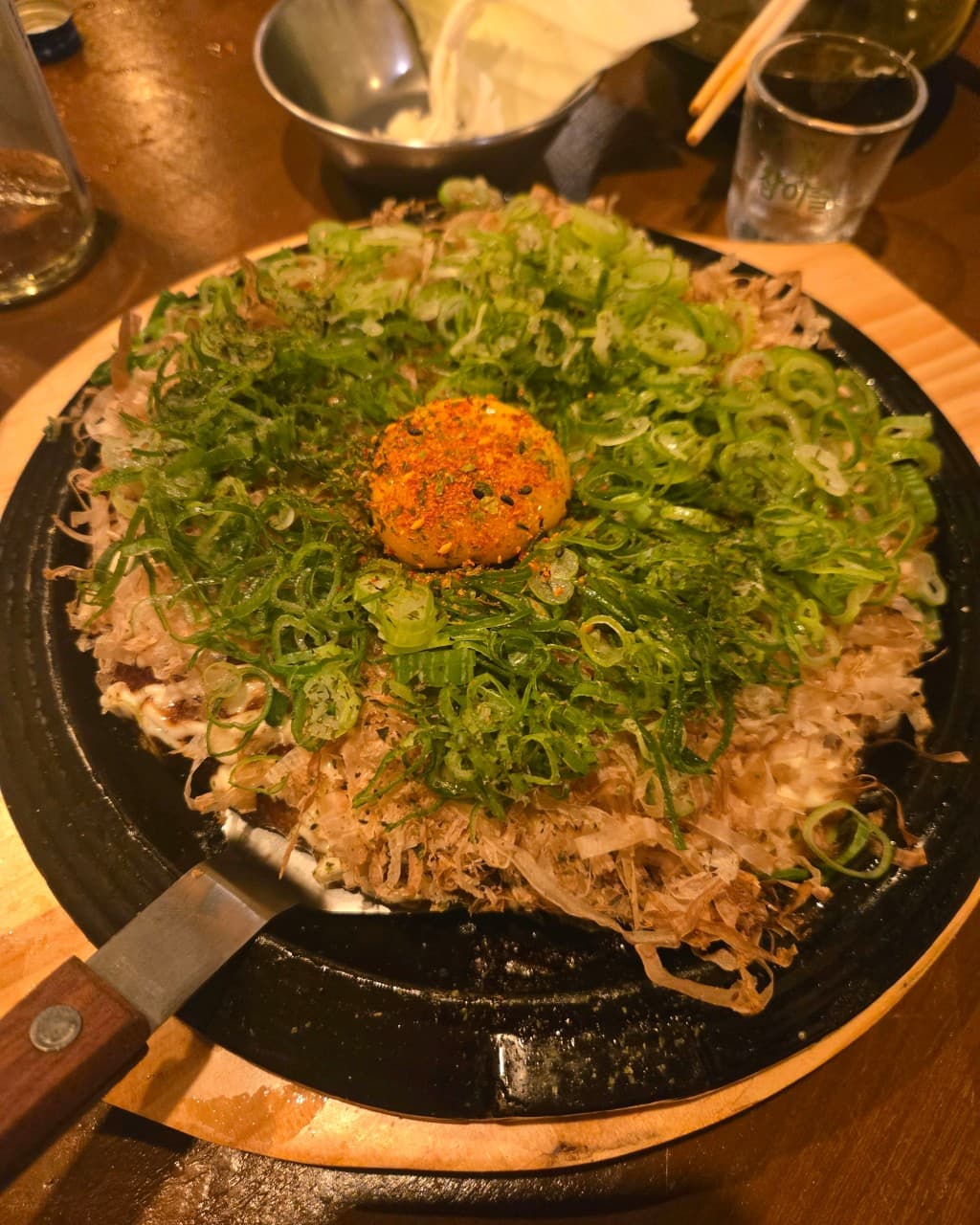 강원특별자치도 양양군 나미야 이자카야 혼술 - 따뜻한 조명 아래 다찌석, 파와 가쓰오부시가 듬뿍 올라간 오코노미야키와 노른자, 소주 한 잔이 차려진 테이블.