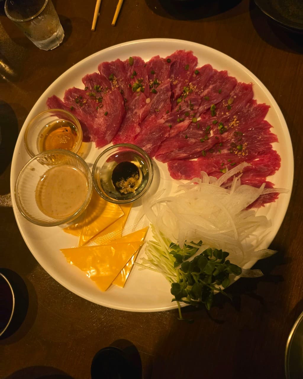 강원특별자치도 양양군 나미야 이자카야 혼술 상황. 어두운 조명 아래 다찌석 위로 선홍빛 육사시미와 치즈, 양파, 소스 세 종류가 담긴 흰 접시가 놓인 1인분 구성.