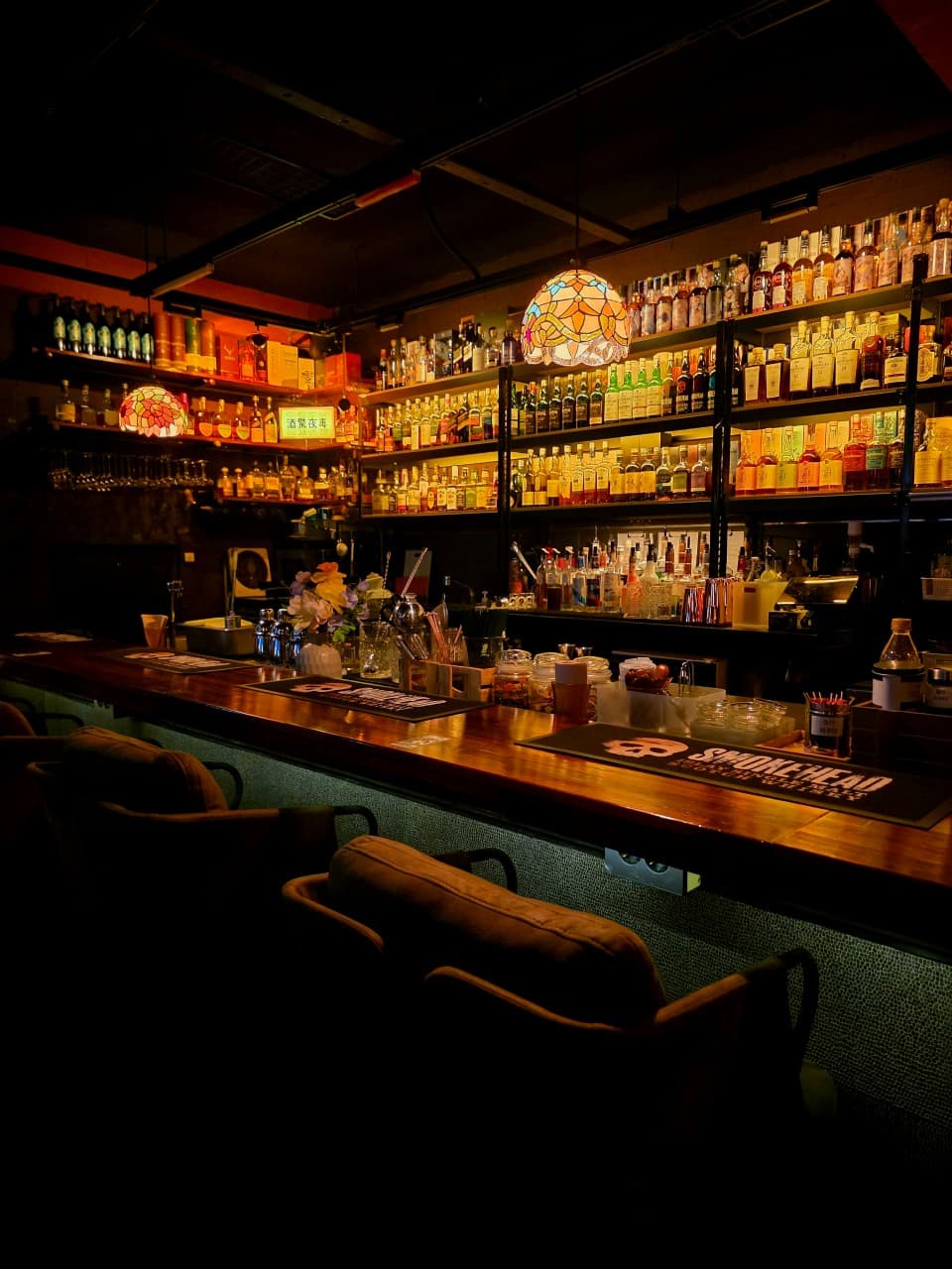 강원특별자치도 속초시 주경야독 바(Bar)의 스테인드글라스 조명이 비추는 긴 원목 다찌석과 1인용 소파. 벽면을 가득 채운 술병들과 혼술을 위한 카운터 공간이 구성됨.