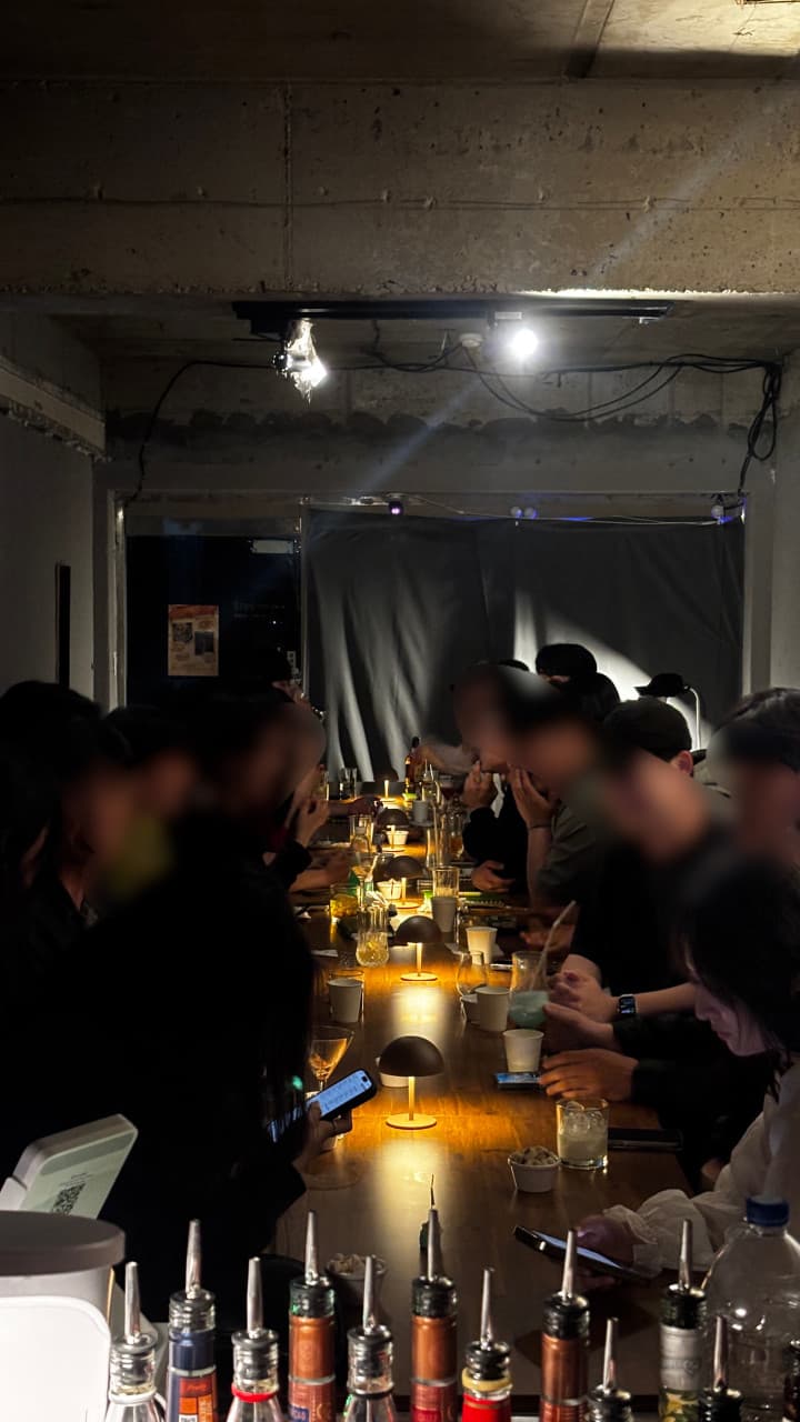 강원특별자치도 강릉시 주인 강릉혼술바 강릉점 바(Bar)의 긴 나무 테이블. 개별 조명 아래 나란히 앉아 칵테일을 즐기는 혼술객들과 다찌석 형태의 실내 전경.