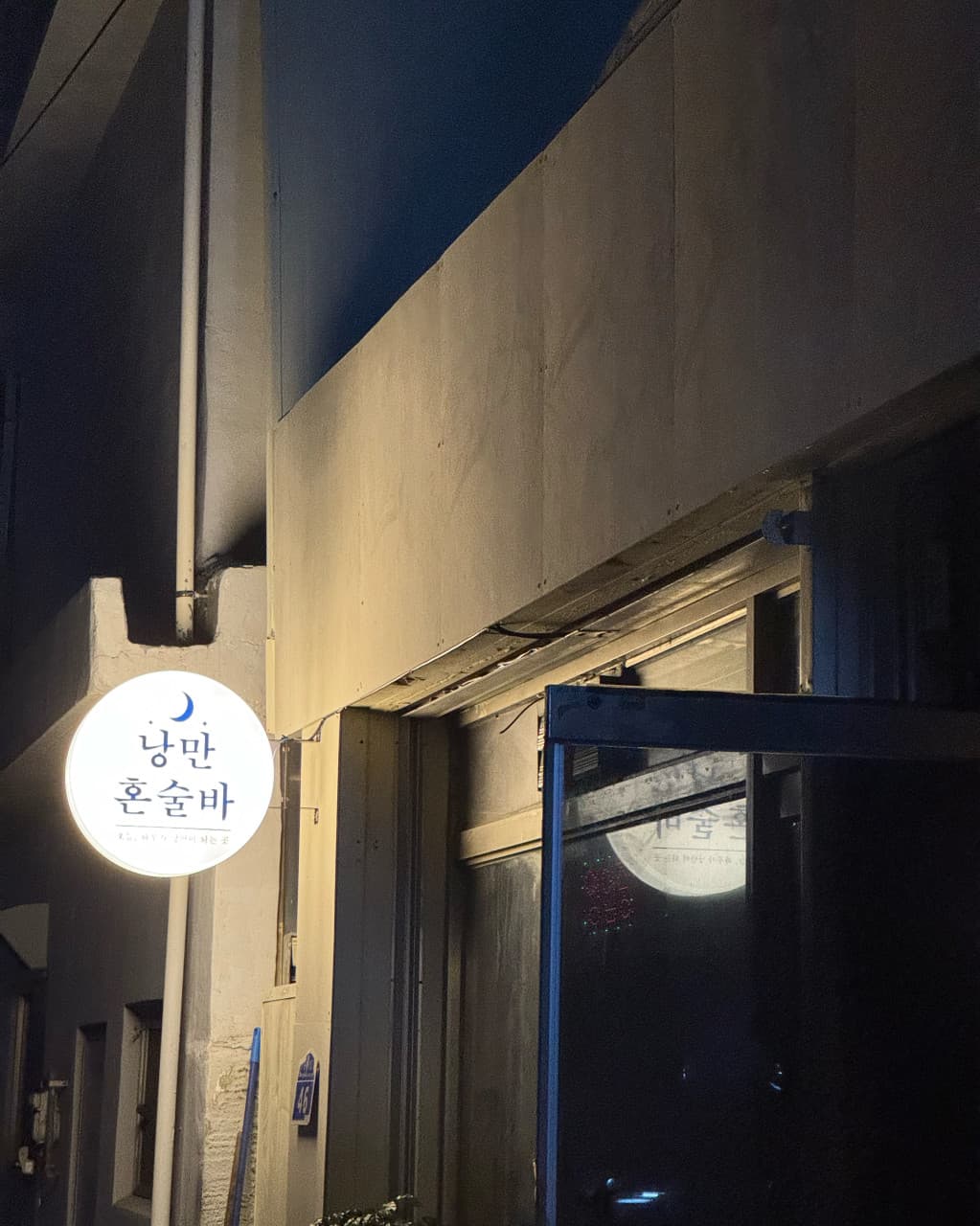 강원특별자치도 강릉시 낭만혼술바 강릉역점 바(Bar) 외관. 밤에 켜진 하얀 원형 간판 조명과 열린 문이 있는 건물 입구. 1인 혼술을 위한 다찌 좌석이 마련된 매장 외부 전경.