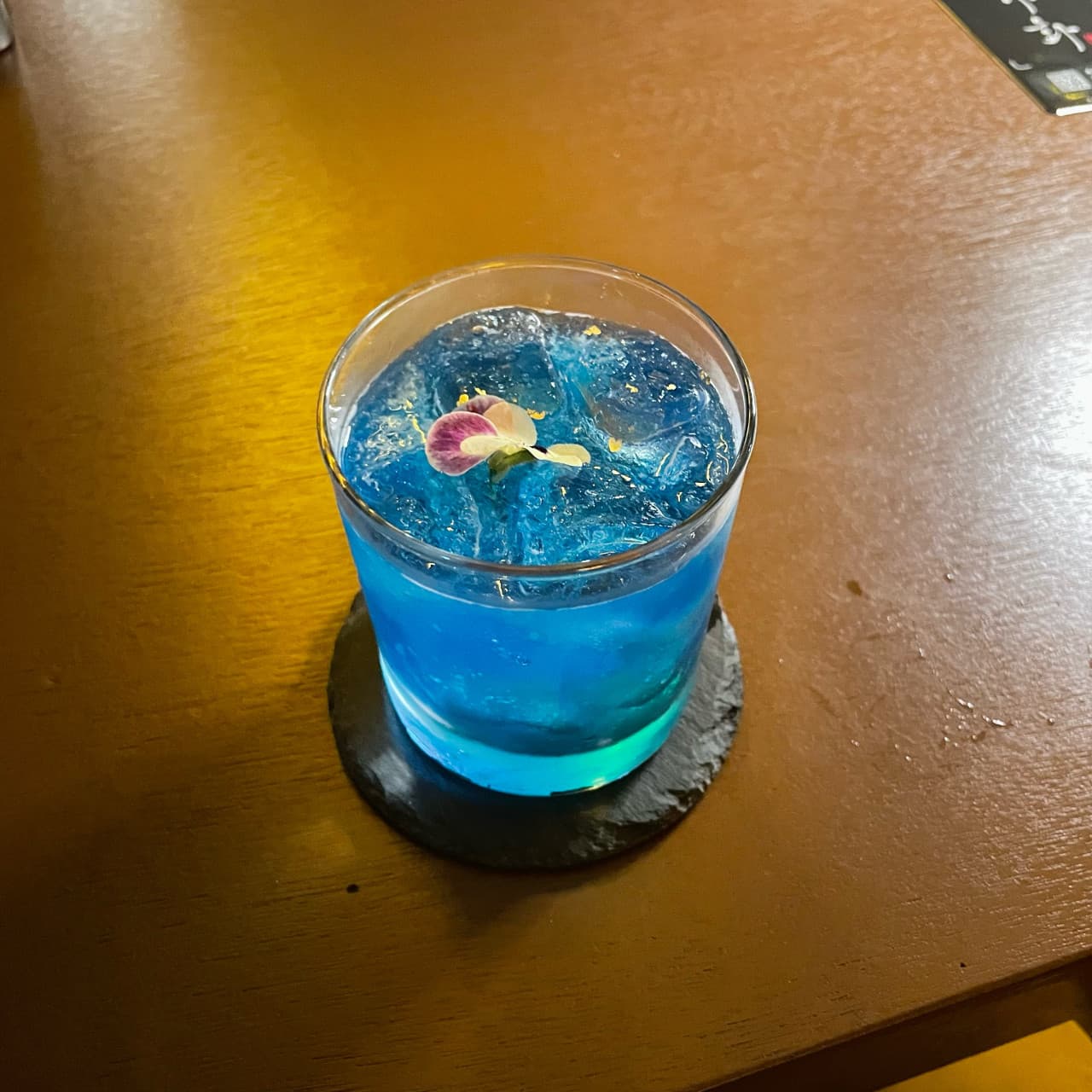 강원특별자치도 강릉시 야화 혼술바 강릉강문해변점 바(Bar) 다찌석에 놓인 파란 칵테일과 식용 꽃. 노란 조명 아래 나무 테이블에서 즐기는 1인 혼술.