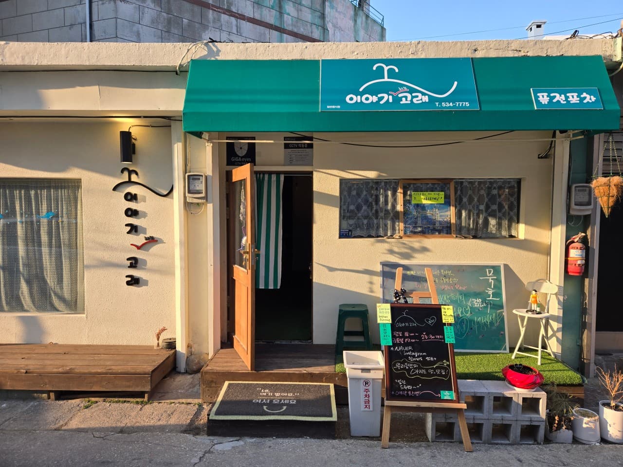 강원특별자치도 동해시 혼술 이자카야 이야기고래 - 청록색 어닝 아래 나무 문과 메뉴 입간판이 배치된 외벽 위로 오후 햇살이 비침