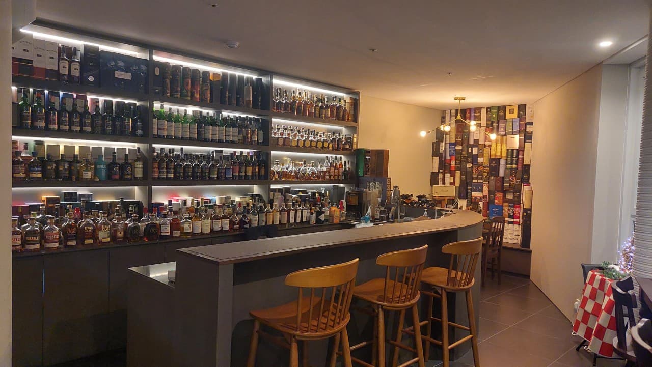 강원특별자치도 속초시 카디어스 바(Bar)의 간접 조명이 비치는 다찌석과 벽면을 가득 채운 위스키병. 혼술을 위해 마련된 원목 의자와 긴 카운터석이 배치된 내부.