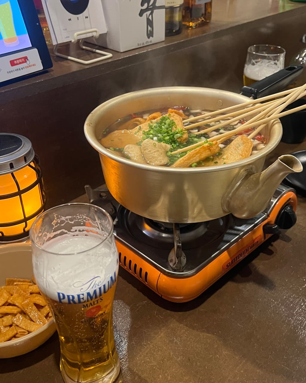 강원특별자치도 원주시 육지놈들 오뎅바 다찌석 혼술. 황색 조명 아래 김이 나는 오뎅 꼬치 전골과 거품 덮인 생맥주가 차려진 나무 카운터석.
