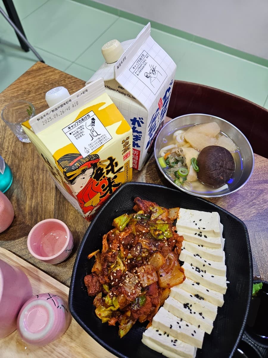 강원특별자치도 속초시 오뎅하우스 오뎅바 - 다찌석 1인 혼술 상차림으로 조명 아래 팩사케와 표고버섯 오뎅탕, 두부김치의 질감이 나무 테이블에 선명하게 드러남.