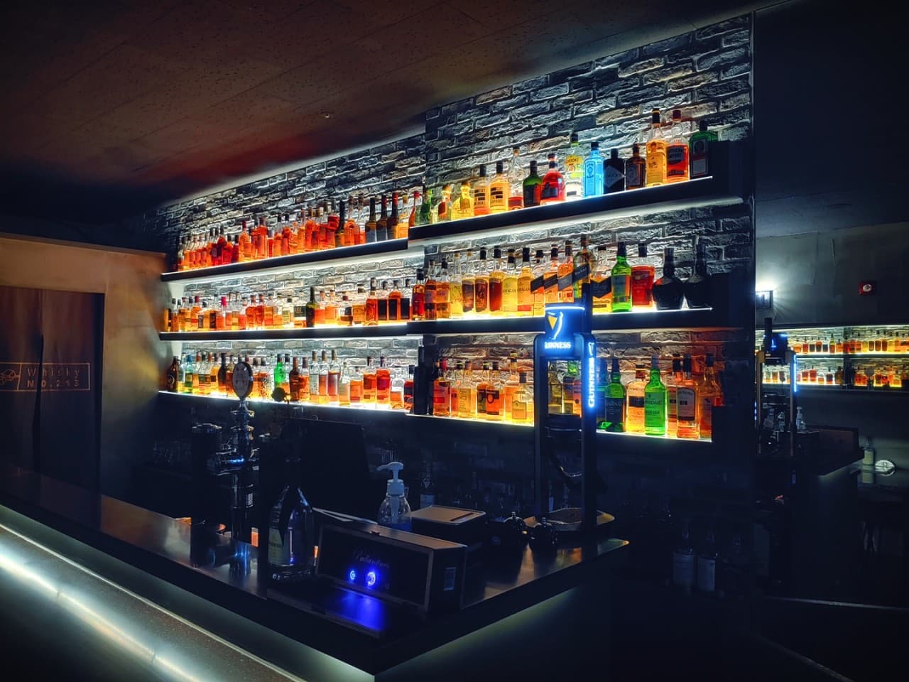강원특별자치도 춘천시 Whisky no.213 바(Bar) 조명 선반에 진열된 위스키 병들과 다찌석. 어두운 공간 속 빛나는 술병들이 배치된 카운터 테이블에서 즐기는 1인 혼술 상황.