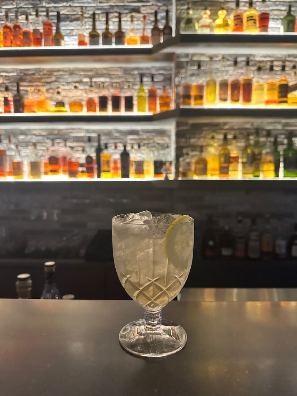 강원특별자치도 춘천시 Whisky no.213 바(Bar) 다찌석 위 얼음과 레몬이 담긴 하이볼. 백라이트 조명을 받은 위스키 진열장 앞 1인 혼술 상황 묘사.