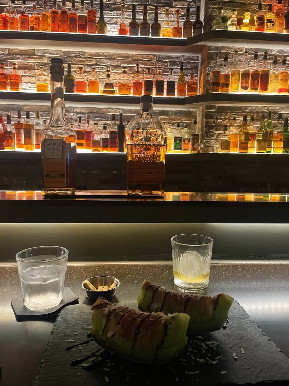 강원특별자치도 춘천시 Whisky no.213 바(Bar) 다찌석 1인 혼술. 조명 비치는 선반 앞 멜론 프로슈토와 얼음 담긴 위스키 잔, 다양한 술병이 나열된 배경