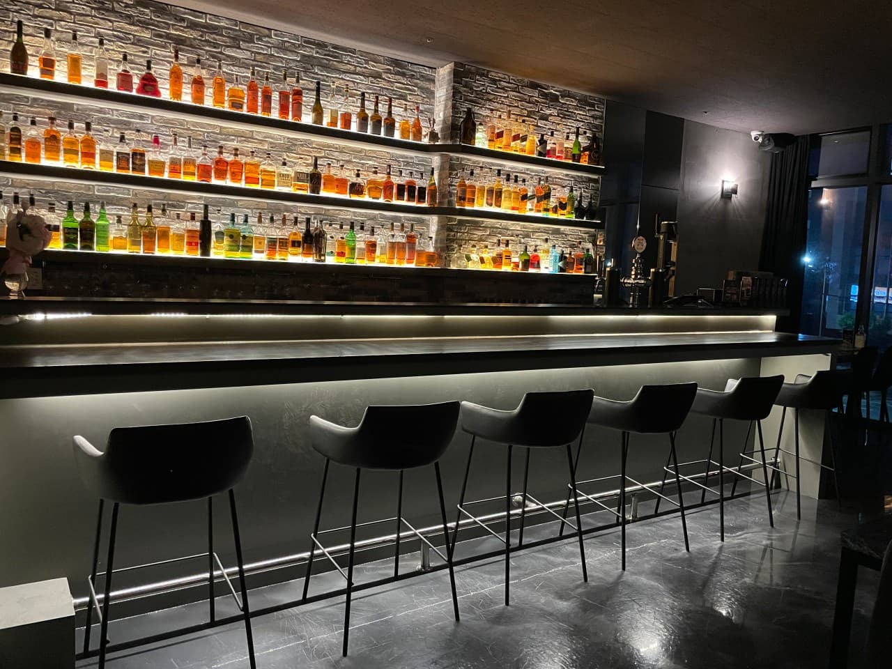 강원특별자치도 춘천시 Whisky no.213 바(Bar)의 간접 조명이 비치는 긴 다찌석과 벽면 가득 진열된 위스키 병. 1인 혼술을 위해 일렬로 배치된 높은 의자와 어두운 실내 구성.