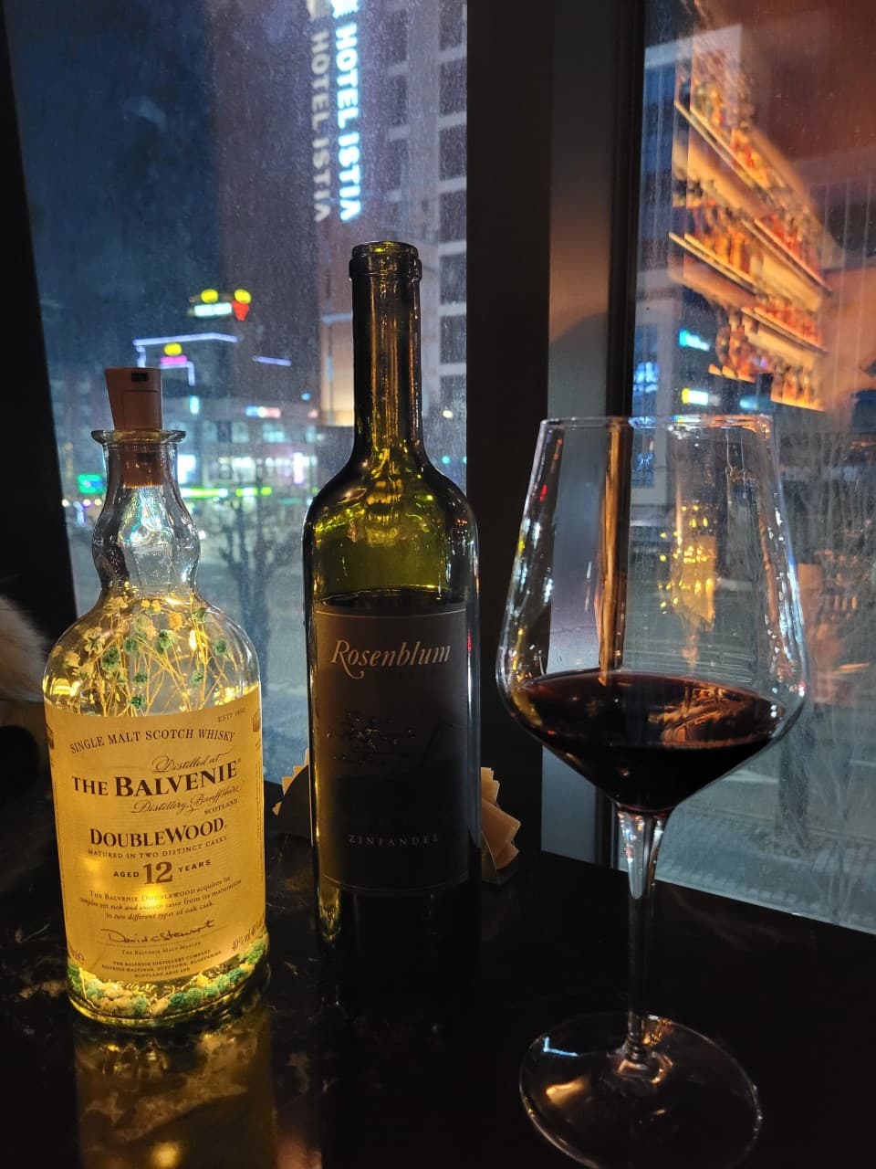 강원특별자치도 춘천시 Whisky no.213 바(Bar) 창가 1인석. 조명이 켜진 위스키 병과 레드 와인 한 잔 너머로 도시 야경이 비치는 혼술 상황.