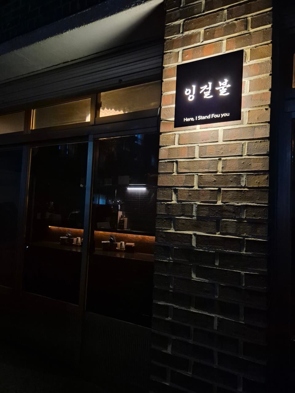 강원특별자치도 강릉시 잉걸불 - 붉은 벽돌 외벽과 창 너머 보이는 1인 다찌석. 노란 조명이 비치는 바 테이블 형태의 혼술 전문 기타 주점.