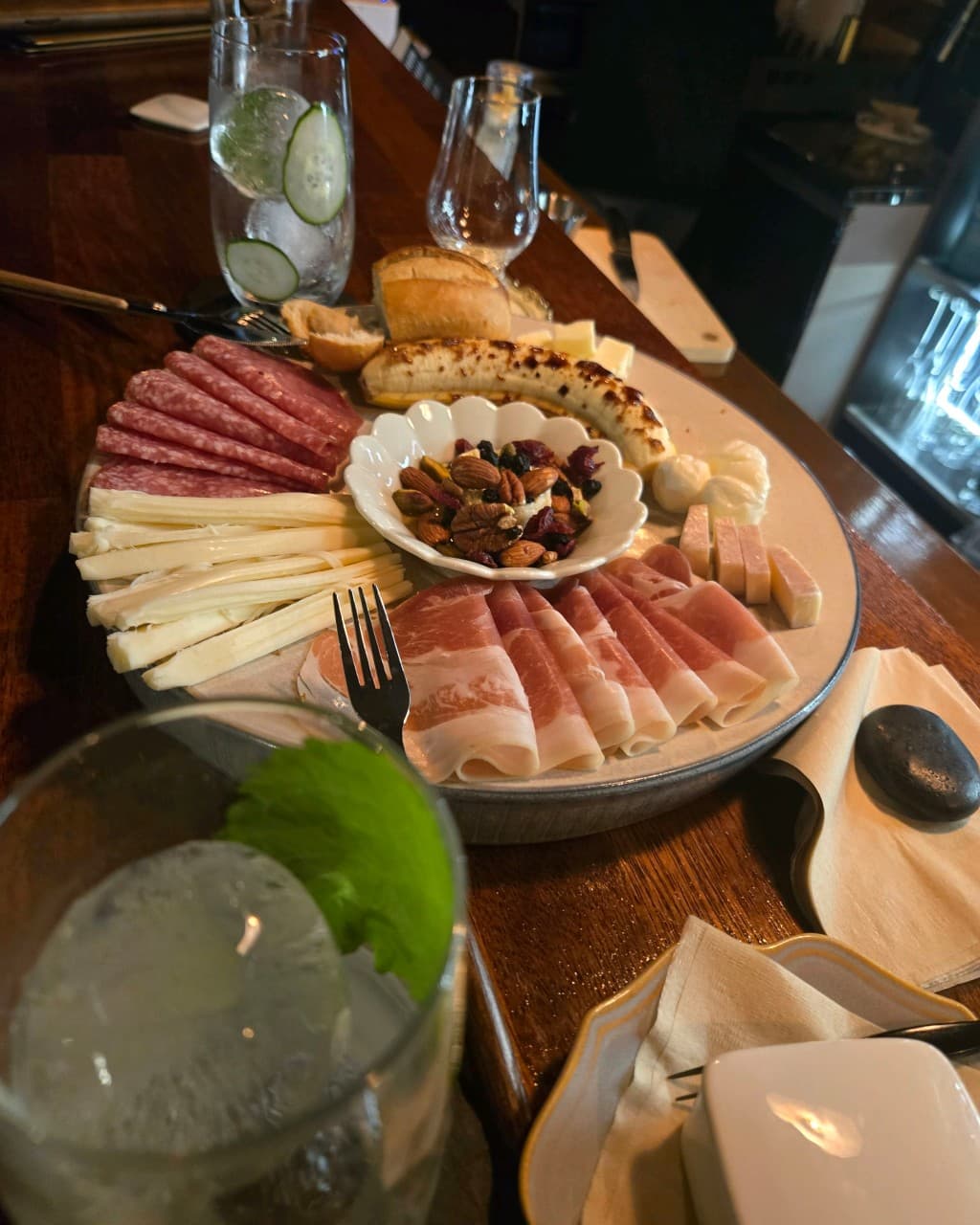 강원특별자치도 철원군 쁘띠클로젯 사사로운공간 바(Bar) 혼술 - 나무 다찌석 위 하몽, 치즈, 견과류 플래터와 하이볼. 1인 좌석에 비치는 조명과 식재료의 질감이 강조됨.