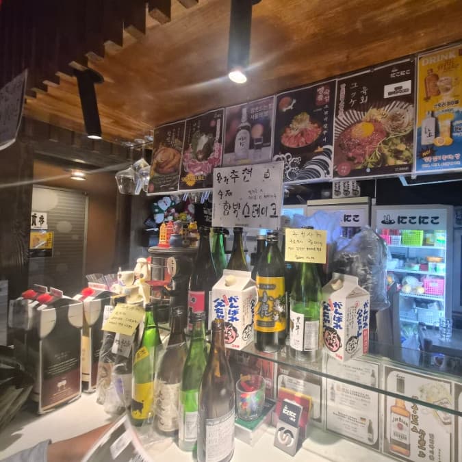 강원특별자치도 영월군 이자카야 니코니코 다찌석. 상단 조명이 비추는 카운터에 나열된 사케 병들과 메뉴 포스터가 어우러진 1인 혼술 공간.