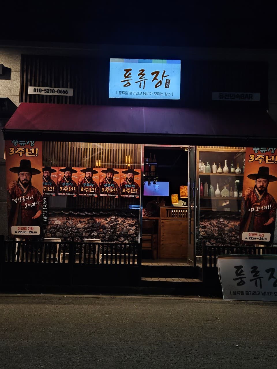 강원특별자치도 춘천시 전통주점 풍류장 입구. 야간 조명 아래 목재 다찌석이 보이는 개방된 문과 한복 포스터가 부착된 돌담 외벽으로 혼술 상황을 표현함.