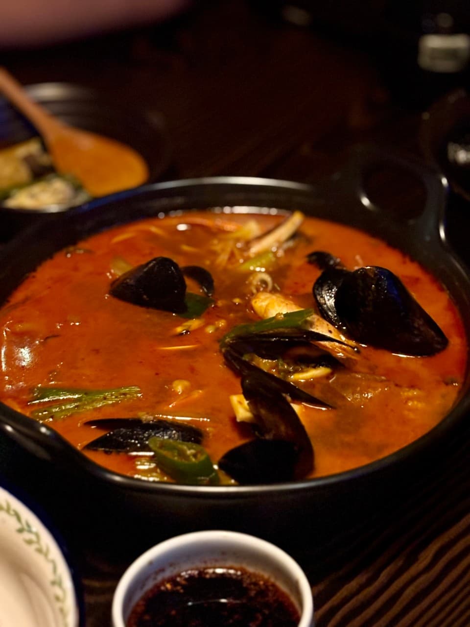 강원특별자치도 춘천시 전통주점 풍류장 다찌석에 차려진 홍합탕. 낮은 조도의 조명 아래 어두운 목재 테이블에서 혼술을 즐기는 상황이며 붉은 국물과 해산물의 질감이 선명함