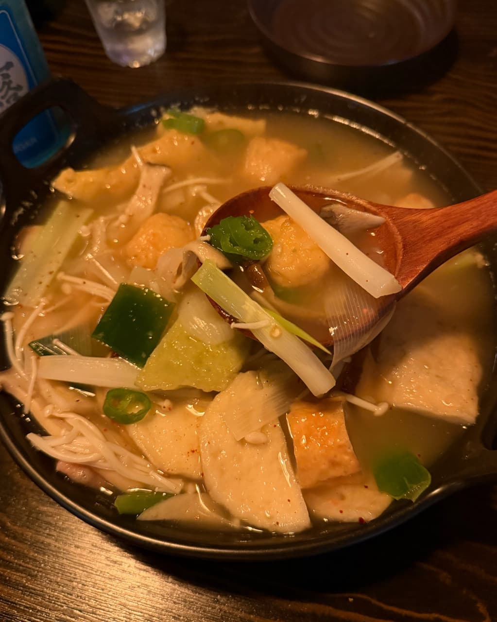 강원특별자치도 춘천시 전통주점 풍류장 다찌석 조명 아래 대파와 팽이버섯을 곁들인 어묵탕 1인 혼술. 목재 숟가락으로 건더기를 뜬 질감과 어두운 나무 테이블 위 차림새.