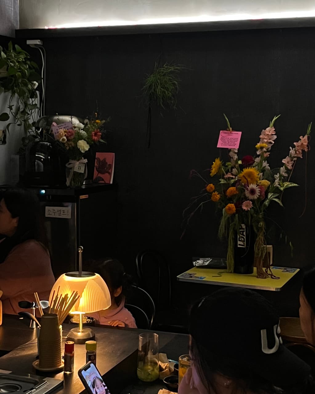 강원특별자치도 춘천시 감마샵앤바 기타 공간 - 노란 스탠드 조명 아래 1인 다찌석과 꽃병이 놓인 어두운 실내. 하이볼 잔이 배치된 혼술 상황