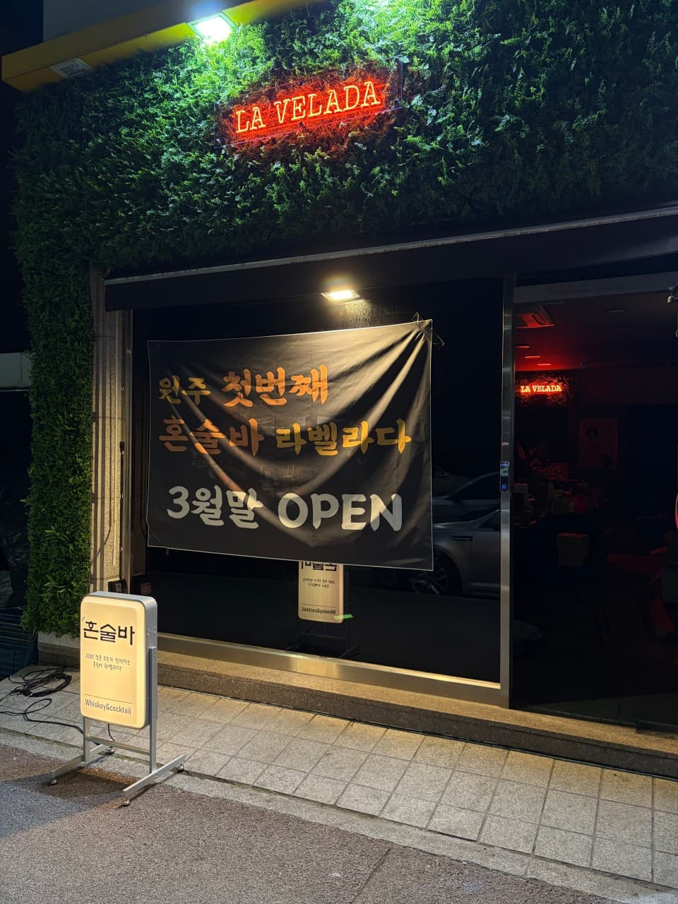 강원특별자치도 원주시 혼술 바 라벨라다 입구. 붉은 네온사인과 초록색 수풀 장식 아래 오픈 현수막이 걸려 있으며 내부 다찌석의 붉은 조명이 보이는 상황.
