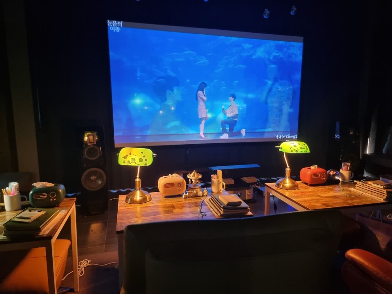 강원특별자치도 강릉시 강릉골방 와인 - 대형 스크린 앞 초록색 조명이 켜진 1인 다찌석에서 혼술하며 영상을 시청하는 목재 테이블과 필기구가 비치된 내부 상황
