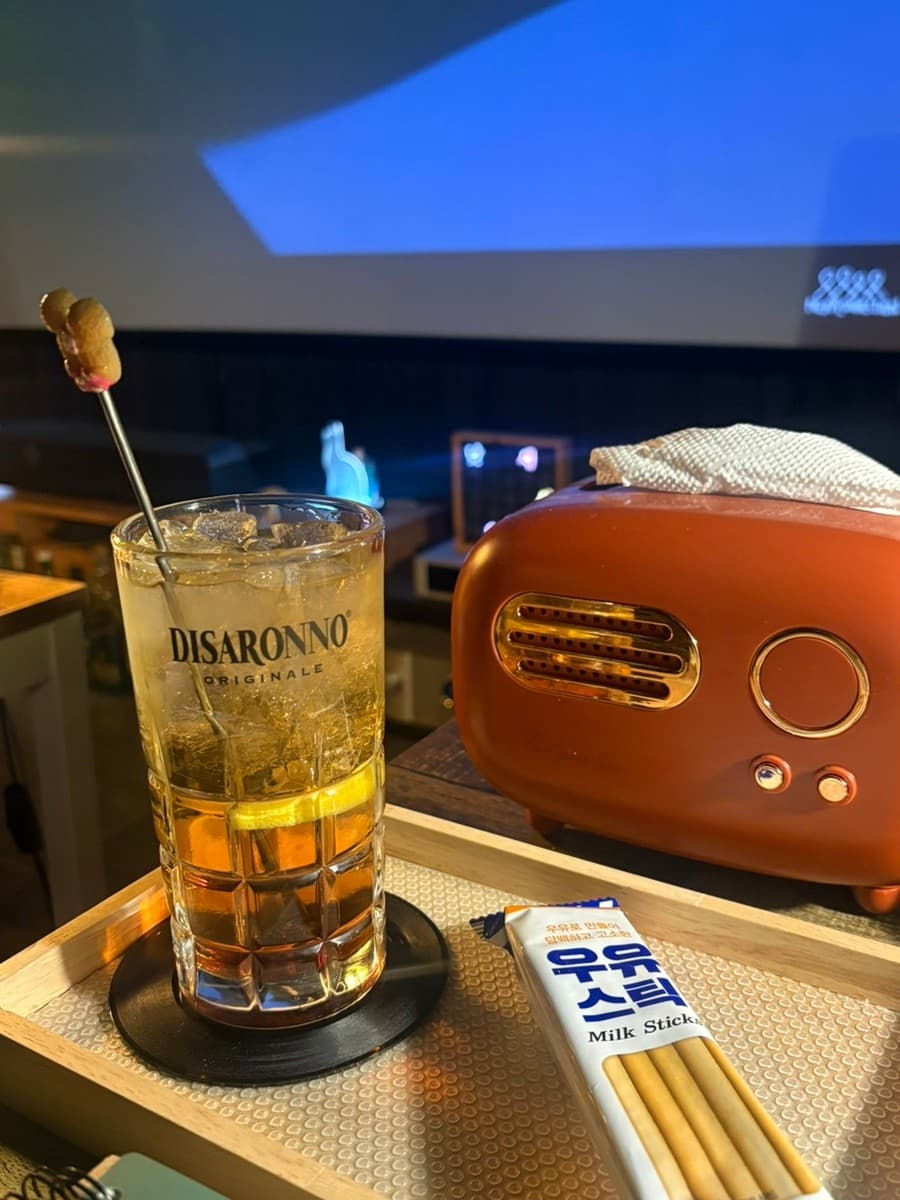 강원특별자치도 강릉시 강릉골방 와인 바 1인 다찌석. 어두운 조명 아래 대형 스크린과 주황색 레트로 소품, 디사론노 하이볼과 우유스틱이 차려진 혼술 상황.