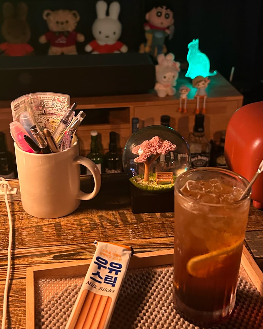 강원특별자치도 강릉시 강릉골방 와인 바 다찌석에서 즐기는 혼술. 어두운 조명 아래 얼음 가득한 잔과 우유스틱, 캐릭터 인형이 배치된 1인석 테이블 공간.