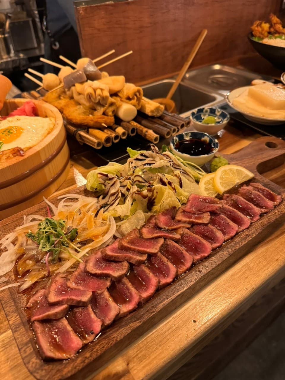 강원특별자치도 원주시 요리주점 철길부산집 원주단계점 다찌석 혼술. 주황빛 조명 아래 선분홍색 소고기 타다끼와 어묵 꼬치가 차려진 1인용 나무 테이블 상황.