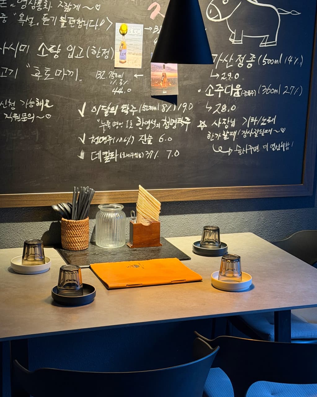 강원특별자치도 강릉시 요리주점 오조의 핀 조명 아래 배치된 1인 식기와 메뉴판. 손글씨 메뉴 칠판 앞 좌석에서 혼술 상황을 연출하며 어두운 내부와 대비되는 테이블 묘사.
