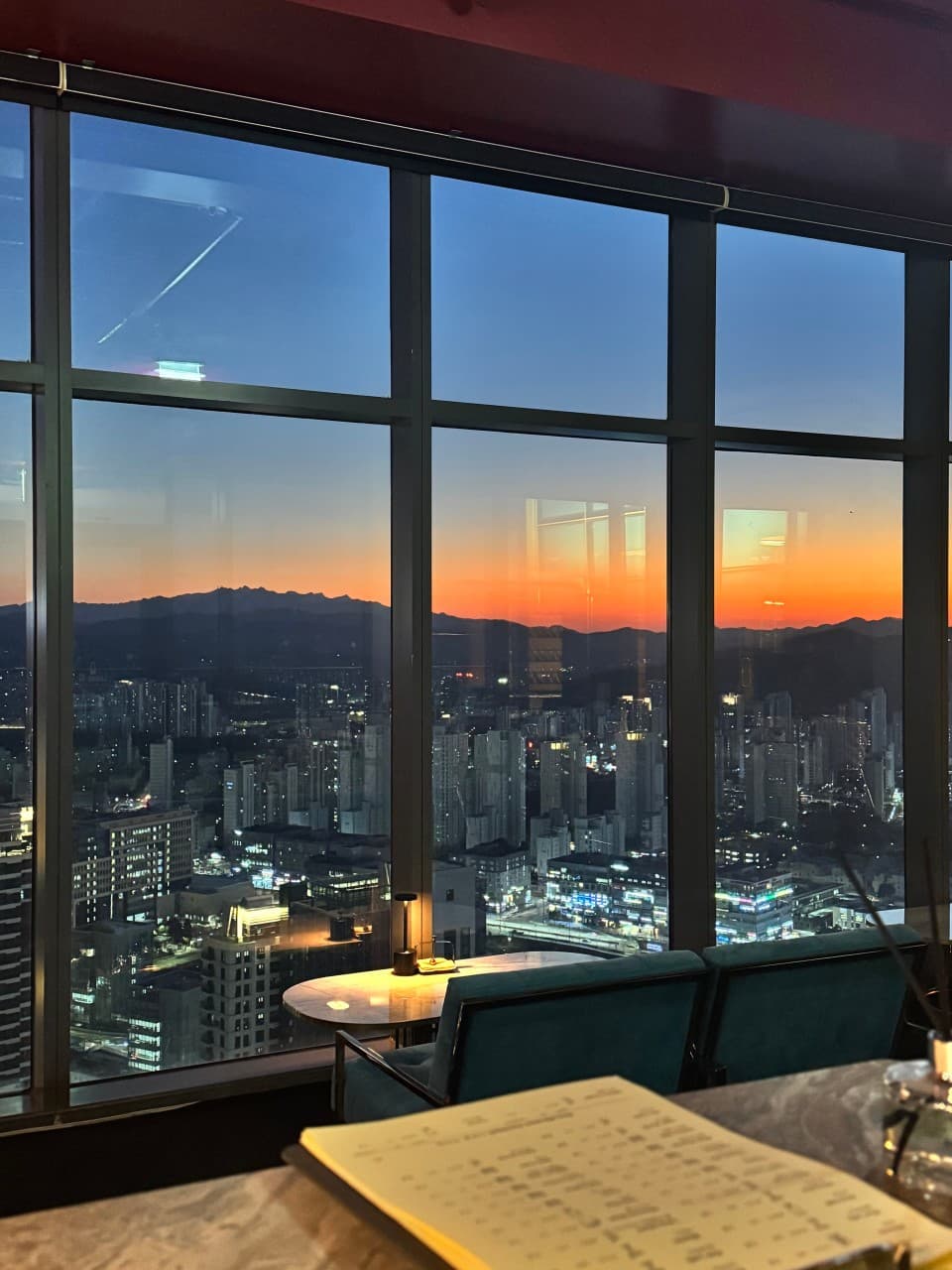 세종특별자치시 나성북1로 라운지46 창가 1인석에서 보이는 주황빛 노을과 도시 전경. 테이블 위 작은 조명과 청색 소파가 놓인 혼술 분위기의 기타 공간.