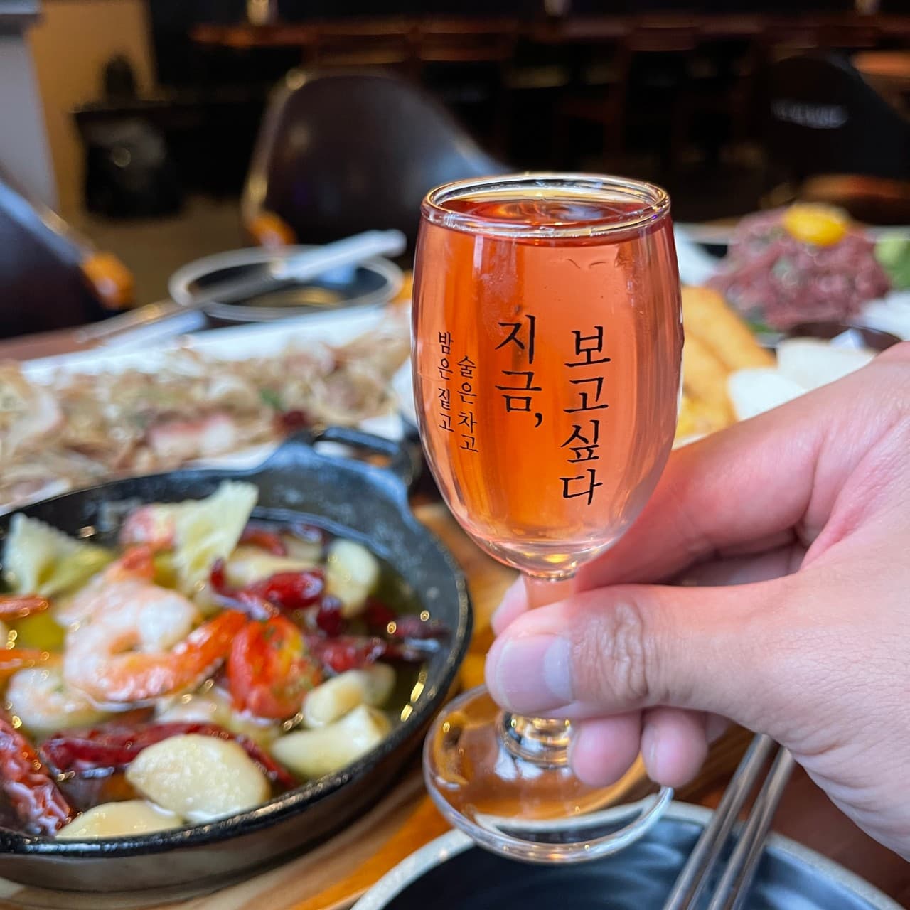 세종특별자치시 나성로 요리주점 지금보고싶다 세종나성점 1인 다찌석. 감바스 알 아히요와 붉은 술이 담긴 잔을 든 손. 어두운 조명 아래 혼술을 하는 상황.