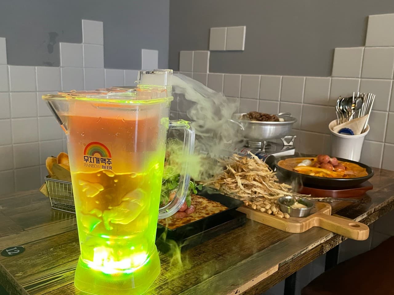 세종특별자치시 해밀3로 무지개맥주 세종해밀점 맥주 호프. 녹색 조명과 연기가 흐르는 맥주 피처, 바삭한 먹태와 떡볶이가 차려진 1인 다찌석에서 혼술을 즐기는 상황.