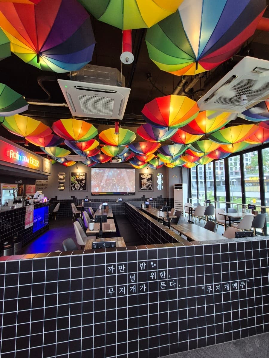 세종특별자치시 해밀3로 무지개맥주 세종해밀점 맥주 호프. 천장 가득한 무지개 우산과 검은 타일 파티션이 있는 1인 혼술 가능 좌석. 대형 스크린과 창가 테이블이 배치된 내부 인테리어 구성.