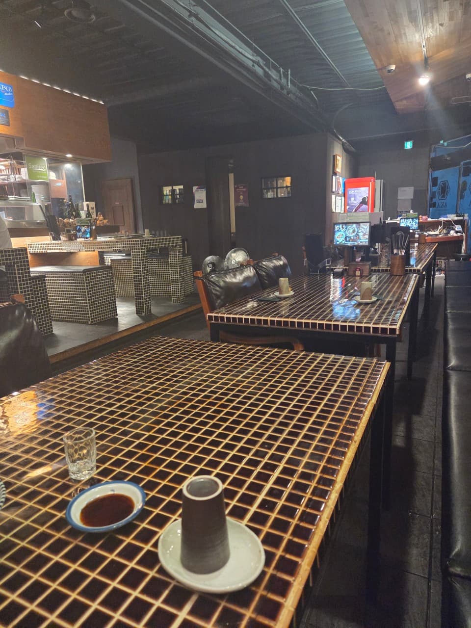 경상남도 양산시 이자카야 아카의 타일 테이블과 1인 다찌석. 낮은 조도 아래 간장 종지와 컵이 놓인 혼술 공간. 세라믹 타일 질감의 가구와 테이블마다 배치된 태블릿 메뉴판.