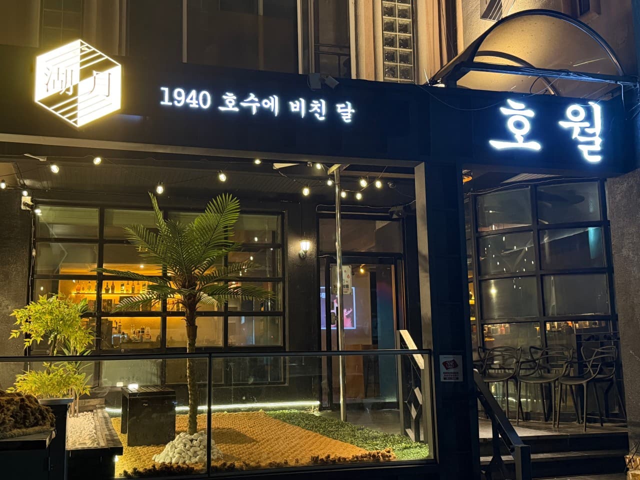 경상남도 진주시 1940호월의 노란 전구 조명과 야자수 조형물이 설치된 매장 입구. 유리창 너머로 혼술 가능한 다찌석과 내부 테이블이 배치된 야간 매장 풍경.