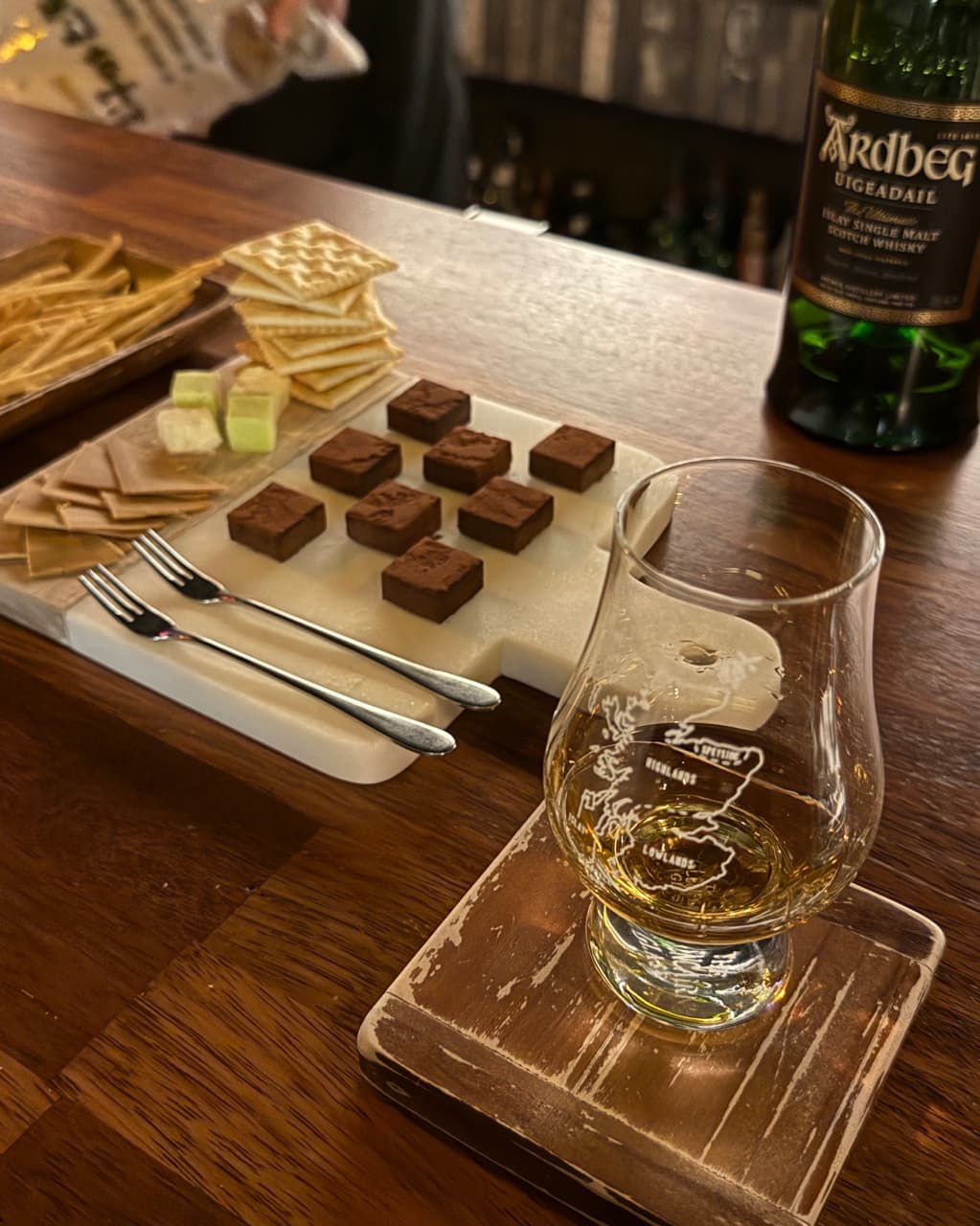 경상남도 창원시 바 보다의 나무 카운터석 위 위스키와 생초콜릿. 어두운 조명 아래 1인 다찌석에서 즐기는 싱글몰트 위스키와 크래커 안주.