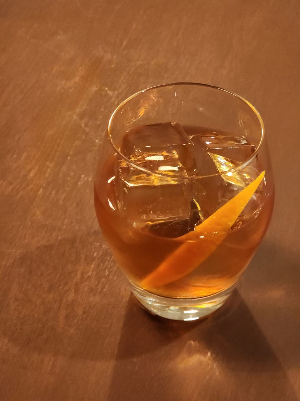 경상남도 진주시 바(Bar) 우심방 나무 테이블 위 혼술용 위스키. 큰 얼음과 오렌지 가니쉬가 담긴 잔 위로 노란 조명이 반사되는 1인 다찌석 상황.
