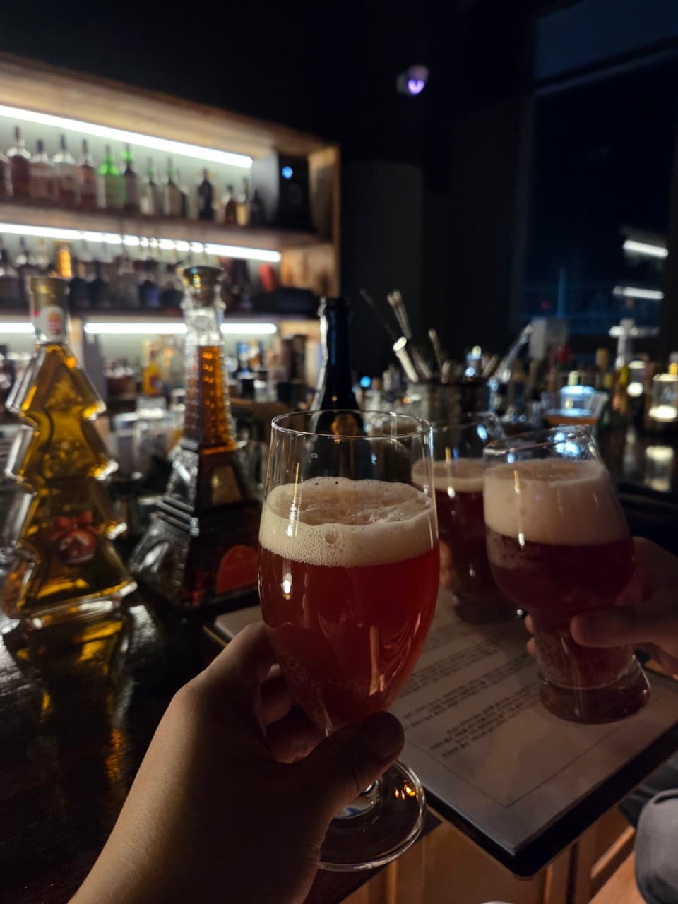 경상남도 진주시 미드나잇덴 바(Bar) 다찌석에서 붉은빛 술과 거품이 담긴 잔을 든 모습. 백라이트 선반에 진열된 술병들과 1인 혼술이 가능한 카운터 공간.
