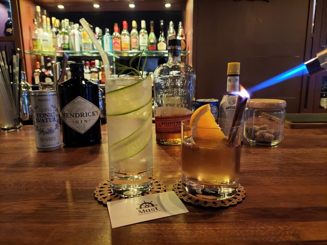 경상남도 창원시 혼술 바(Bar) 마스트 - 나무 다찌석 위 오이 슬라이스 칵테일과 토치 불꽃으로 시나몬을 그을리는 연출. 진열된 술병들과 조명 아래 놓인 술잔.