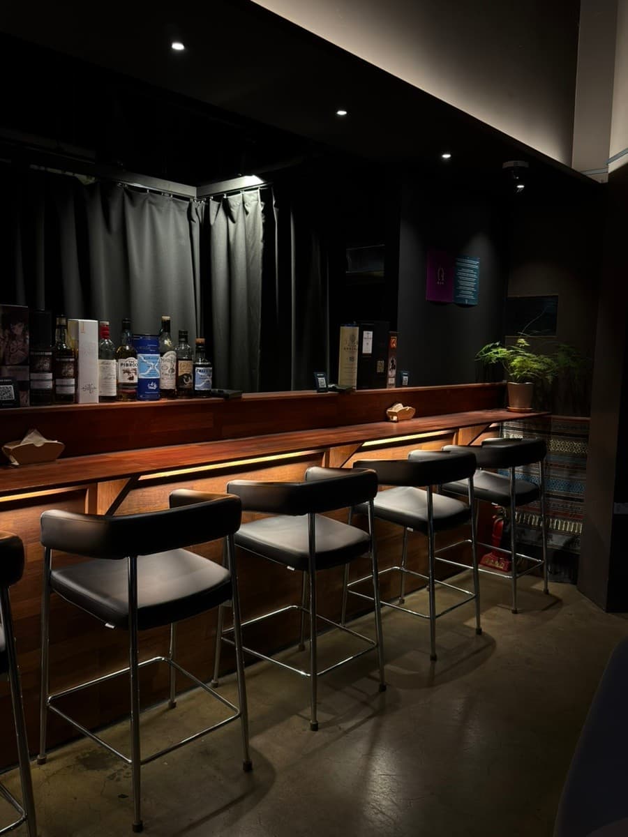 충청북도 청주시 위숙희 바(Bar)의 어두운 조명 아래 놓인 나무 다찌석과 검정색 1인용 의자. 진열된 위스키 병들과 하단 간접 조명이 비치는 카운터에서 혼술 상황을 조성함.