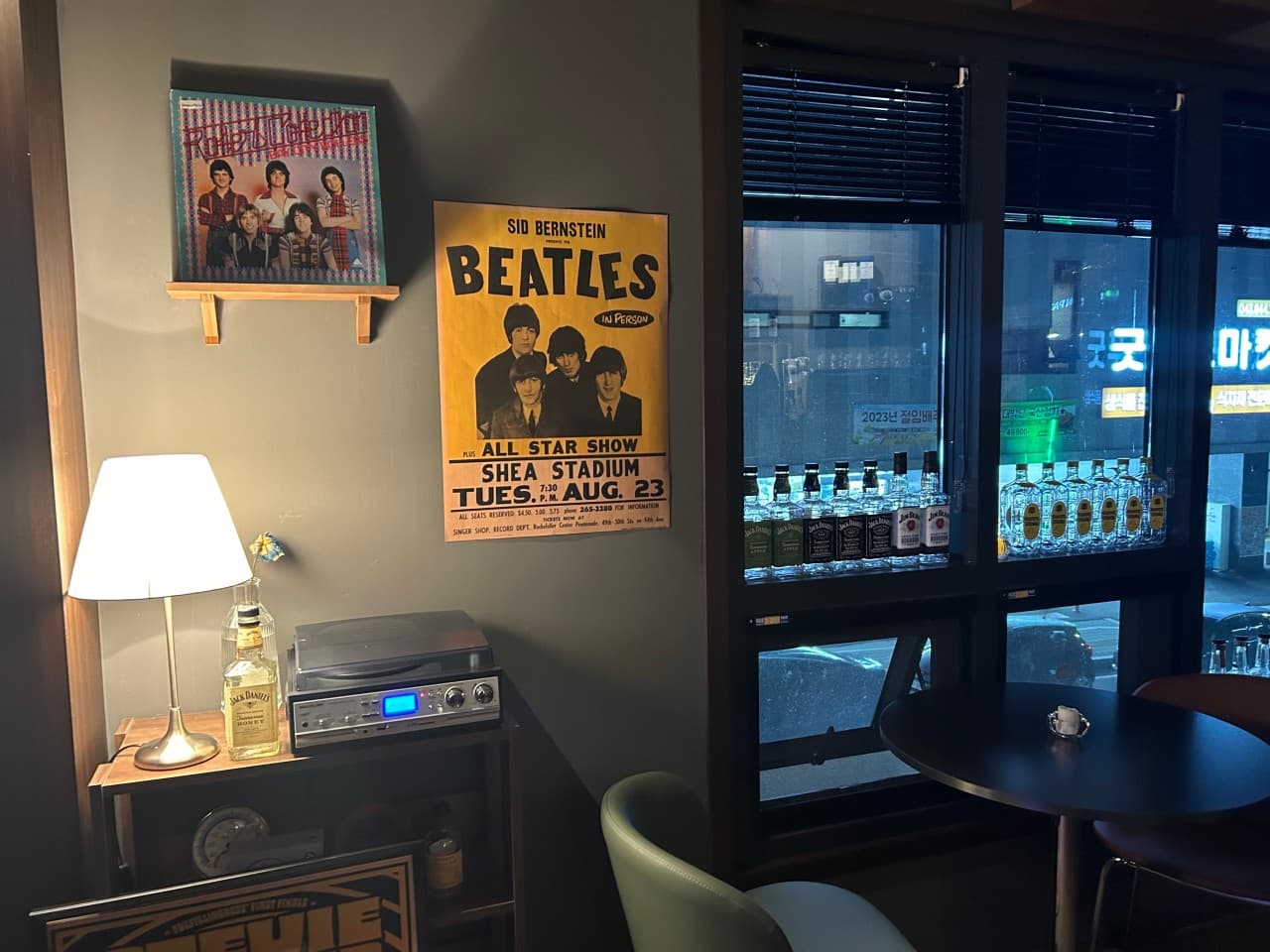 충청북도 충주시 혼술 바(Bar) 조용 - 스탠드 조명과 비틀즈 포스터가 있는 벽면, 위스키 병이 나열된 창가 쪽 1인 원형 테이블과 좌석 구성