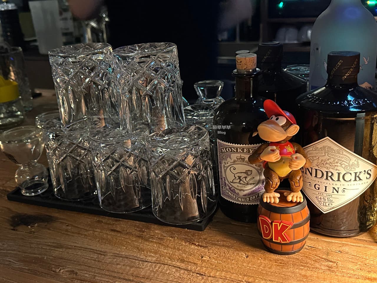 충청북도 충주시 매드슬로우 용산 바(Bar) 다찌석에 진열된 몽키47 진 보틀과 정교한 유리잔들. 은은한 조명 아래 피규어가 놓인 원목 카운터에서 즐기는 1인 혼술 상황.