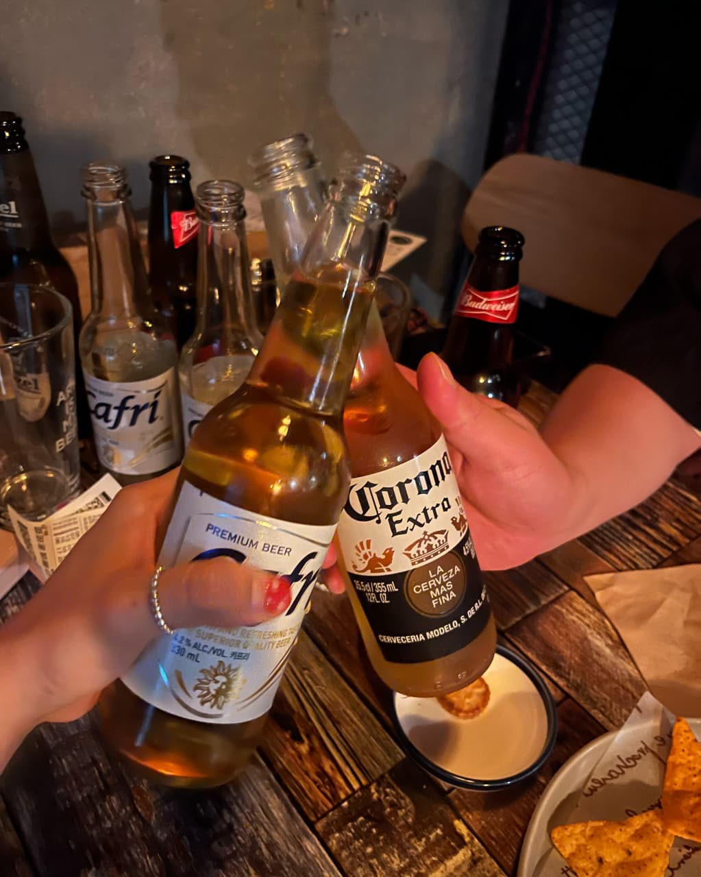 충청북도 충주시 벙커 LP Bar - 조명 아래 나무 테이블에서 카페리와 코로나 병맥주를 부딪치는 상황. 나초와 빈 병이 놓인 1인 다찌석 중심의 LP바 내부