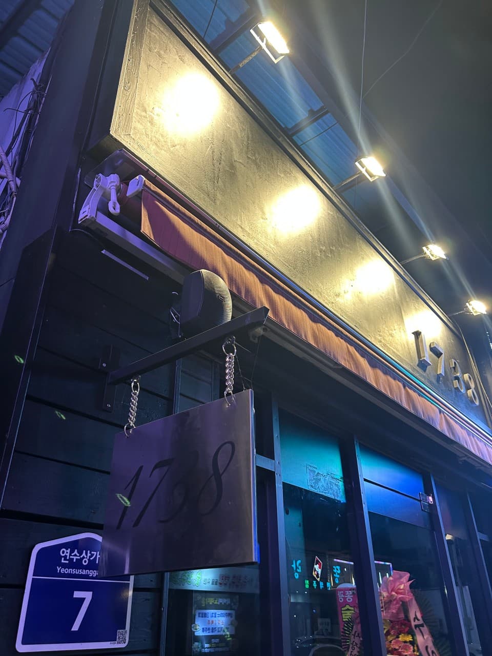 충청북도 충주시 1738pub 연수점 바(Bar)의 야간 외관. 상단 핀 조명이 비추는 금속 간판과 연수상가 주소판이 위치함. 혼술 손님을 위한 어두운 목재 외벽과 보랏빛 조명이 대비되는 입구.