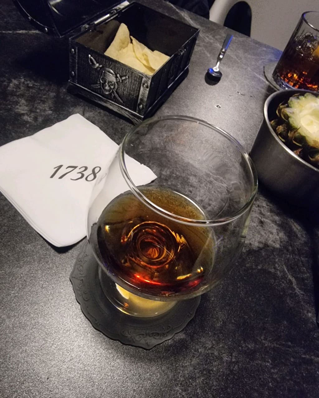충청북도 충주시 1738pub 연수점 바(Bar)의 혼술 차림. 어두운 테이블 위 1738 로고 냅킨과 브랜디 잔, 보물상자 속 나초와 파인애플 안주가 놓임.